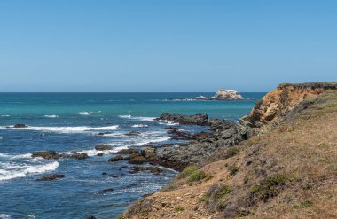 San Simeon, CA, USA - 8 Haziran 2021: Şehrin kuzeyinde Pasifik Okyanusu kıyısı. Mavi-gök mavisi okyanustaki Piedras Blankas adası ve beyaz sörflü alçak kahverengi ve siyah uçurumlar.
