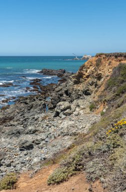 San Simeon, CA, USA - 8 Haziran 2021: Şehrin kuzeyinde Pasifik Okyanusu kıyısı. Mavi-mavi okyanustaki Piedras Blankas adasının portresi ve beyaz sörflü açık kahverengi ve siyah uçurumlar.