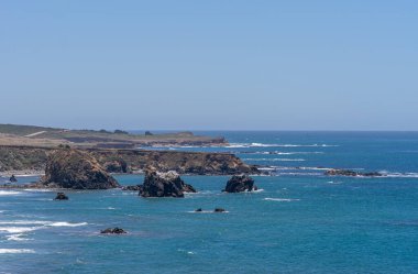San Simeon, CA, ABD - 8 Haziran 2021: Şehrin kuzeyinde Pasifik Okyanusu kıyısı. Açık mavi gökyüzünün altındaki derin mavi sularda siyah kayalıklarla ve kayalarla kaplı bir manzara. Beyaz sörf renk katar.