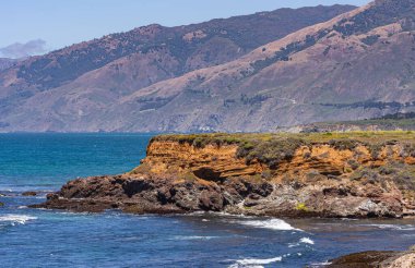 San Simeon, CA, ABD - 8 Haziran 2021: Şehrin kuzeyinde Pasifik Okyanusu kıyısı. Ragged Point yakınlarındaki derin mavi sularda Kızıl kaya kayalıklarının klonlanması. Los Padres dağları arkada..