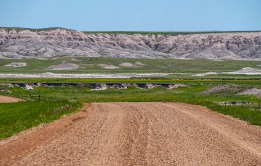 Badlands Ulusal Parkı, SD, ABD - 1 Haziran 2008: Kahverengi çakıl ve toprak yol, mavi gökyüzü altında gri ve beyaz jeolojik çökeltilerini açığa çıkaran tepe sırtlı yeşil çayıra iniyor..