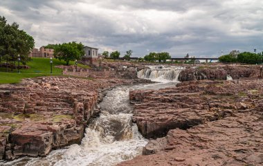 Sioux Falls, SD, USA - June 2, 2008: The main flow of the white foaming water cascade over and between brown rocks under heavy cloudscape with green foliage belt on horizon.