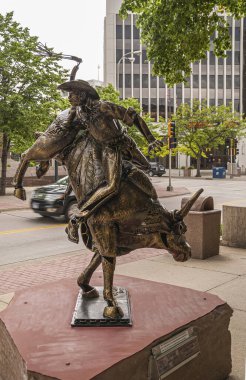 Sioux Falls, SD, USA - June 2, 2008: Closeup of cowboy riding bull bronze statue, titled, Is This Love, along main street with green foliage as backdrop. Street scene with traffic lights.