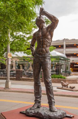 Sioux Falls, SD, USA - June 2, 2008: Closeup of half-naked muscular man drinking from bottle bronze statue along street with parking garage building in back. Some green foliage.