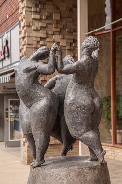 Sioux Falls, SD, USA - June 2, 2008: Closeup of 3 dancing naked obese persons black bronze statue along street with modern style building walls as backdrop.