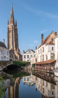 Brugge, Flanders, Belçika - 4 Ağustos 2021: Sunlit Onze Lieve Vrouw Katedrali mavi gökyüzünün altında. Dijver Kanalı 'nın yansıması, sarayların ve evlerin beyaz cepheleriyle Dijver Köprüsü' ne çıkıyor.. 