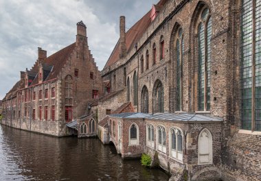 Brugge, Flanders, Belçika - 4 Ağustos 2021: Eski Sint Jans Hastanesi 'nin kahverengi tuğlalı taş cepheleri Mariabrug' daki siyah su kanalı boyunca uzanmaktadır. Gri Bulutlu.