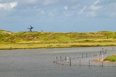 Knokke-Soygunu, Flanders, Belçika - 6 Ağustos 2021: Zwin Doğa Koruma Alanı. Yeşil kumullardaki Siyah Keun heykeli mavi bulutların altında Nord Denizi 'nden tuzlu bataklık deresini ayırıyor.. 