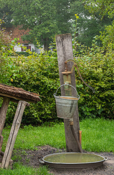 Генк, Бельгия - 11 августа 2021 года: Доменико Боккетти. Крупный план старого зеленоватого водяного насоса, закрепленного на бревне из дерева с металлическим ведром, свисающим с его крана. Зеленая листва.