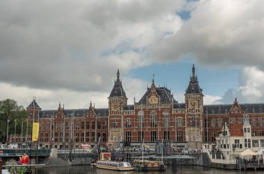 Amsterdam, Hollanda - 14 Ağustos 2021: Kalın mavi bulutlar altında altın süslemeli kırmızı taş Centraal tren istasyonu binası. Turistler su üzerinde tekne kanalı açarlar. Prins Hendrikkade.