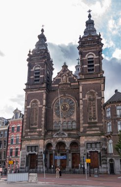 Amsterdam, Hollanda - 14 Ağustos 2021: Gri bulutlar altında süsleri, heykeli ve Saint Nicolas Bazilikası 'nın iki kulesi olan koyu kahverengi heykel. İnsanlarla sokak sahnesi.