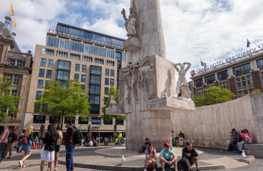 Amsterdam, Hollanda - 14 Ağustos 2021: Ağır mavi bulutlar altında Beyaz Ulusal Anıt. Arkadaki iş binaları ve yeşil yapraklar. Etrafta bir sürü insan var..