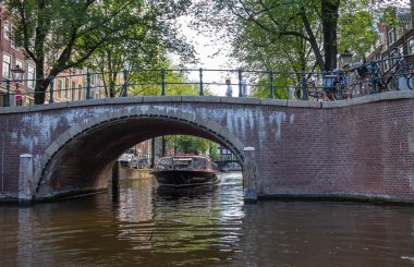 Amsterdam, Hollanda - 15 Ağustos 2021: Kanallarda küçük bir köprü. Yukarıda yeşil yapraklar ve köprü kemerinin altında yolculuğa başlayan bir tekne..