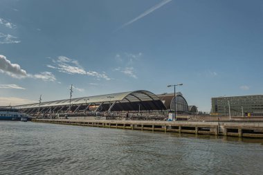 Amsterdam, Hollanda - 15 Ağustos 2021: Centraal İstasyonu üzerinde mavi gökyüzü altında yarım daire cam ve IJ nehir suyu önünde. 