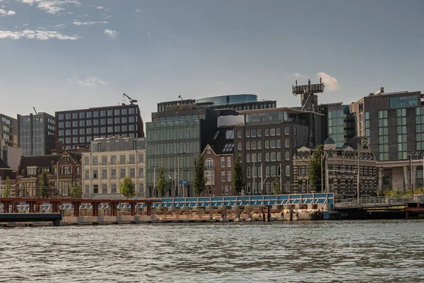 Amsterdam, Hollanda - 15 Ağustos 2021: Centraal İstasyonu 'nun doğusundaki IJ nehri boyunca bulunan eski ve yeni evler ve işyeri binaları açık mavi gökyüzü altında.