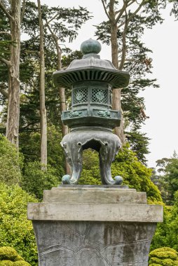 ABD, Kaliforniya, San Francisco - 19 Mayıs 2008: Japon Bahçesi. Yeşil bronz fener heykeli gri taş kaidede, arkasında yeşil yaprakların farklı tonlarında. Gümüş gökyüzü.