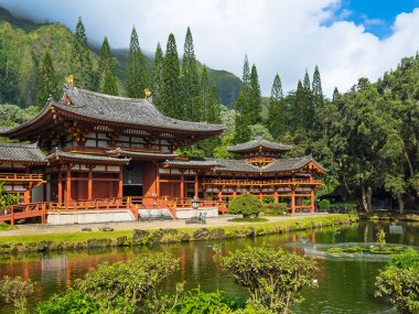 Kaneohe, Oahu, Hawaii, USA. - July 12, 2025: Maroon walls and gray roof, Byodo-In Buddhist temple front facade with koi-pond in front, green trees and mountain range in back under blue cloudscape
