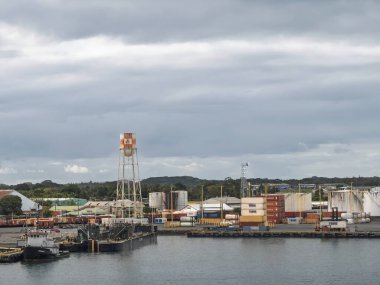 Hilo, Hawaii Island, Hawaii, USA - July 15, 2025: South docks of the harbor with large tanks and tower under light blue cloudscape. Stacks of containers by Young and Pasha Hawaii. Barge on pier