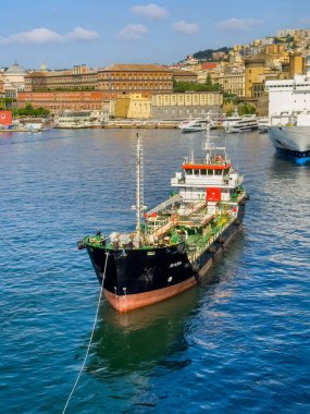 Napoli, İtalya - 17 Temmuz 2024: Liman manzarası. SB Alisea tanker gemisi yaklaşıyor ve yakıt teslimatı için yolcu gemisine bağlanıyor. Kırmızı-kahverengi Enrico Caruso Müzesi ve sarı Parthenope Üniversitesi binaları
