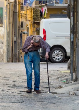 Napoli, İtalya - 17 Temmuz 2024: Bastonlu yaşlı bir adam, 90 derecelik açıyla eğilip, yavaşça sokakta yürürken telefonu kulağına dayadı.