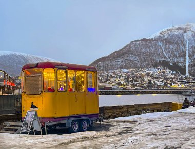 Tromso, Norveç - 23 Aralık 2025: Küçük vagon, anakara dağı ve şehir manzarasıyla birlikte buzun üzerinde, Noel için aydınlatıldı ve dekore edildi.