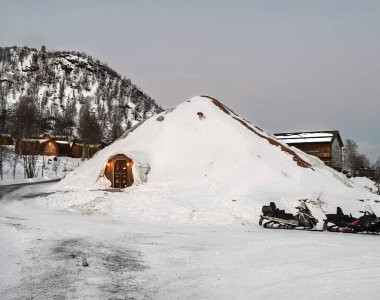 Kirkenes, Norveç - 25 Aralık 2025: Snowhotel 'in karla kaplı büyük igloo bölümünün giriş tarafı. Dağ arkada ve bir grup kar aracı önde.