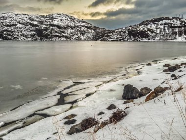 Kirkenes, Norveç - 26 Aralık 2025: Jentoftbukta körfezi çevresindeki doğa, kısmen donmuş. Kıyı şeridinde yıkılmış düzlükler, tepelerin üzerinde gün batımının doğuşu.