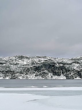 Kirkenes, Norveç - 26 Aralık 2025: Jentoftbukta körfezi çevresindeki doğa, kısmen donmuş. Arka taraftaki dağ sırası ve kalın gri bulutların altında küçük buzlar yüzüyor.