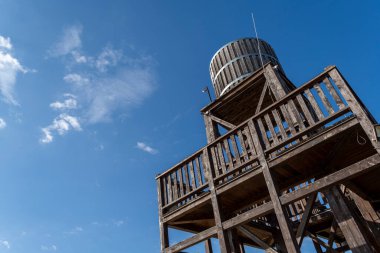 Uzun, ahşap bir su kulesi açık mavi gökyüzüne karşı belirgin bir şekilde duruyor. Açık bir güverteye sahiptir ve huzurlu bir manzaraya sahip kırsal bir alanda yer almaktadır..