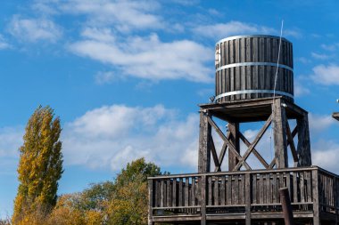 Tahta bir su kulesi renkli sonbahar ağaçlarıyla çevrili ahşap bir platformun üzerinde yükseliyor. Açık mavi gökyüzü ve kabarık bulutlar bu kırsal bölgede sakin ve pitoresk bir manzara yaratır..