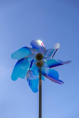 Canlı mavi bir fırıldak rüzgarda zarif bir şekilde dönüyor, parlak mavi bir gökyüzüne karşı. Güneş ışığı parlak yüzeyinden yansıyarak neşeli görünümüne katar..