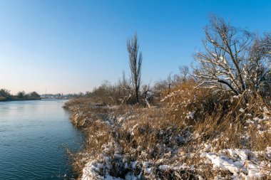 Bir nehir açık mavi bir gökyüzünün altında pürüzsüz bir şekilde akar. Ağaçlar kıyıya dizilir, bazıları karla doludur ve kuru otlar serin havada hafifçe sallanır. Sahne kışın sessiz güzelliğini gösteriyor..