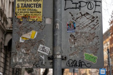 Yıpranmış çıkartmalar ve graffitilerle dolu kentsel bir alanda bir tabela duruyor. Bilgi kısmen gizli. Sahne, şehirde işlek bir caddenin karakterini yakalar..