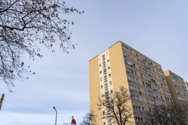 Birkaç bulutu olan açık bir gökyüzüne karşı yüksek bir bina görülür. Yakınlarda ağaçlar boş ve aşağıda bir kaldırım var. Sahne, sıradan bir günde şehir hayatının belirtilerini gösteriyor..