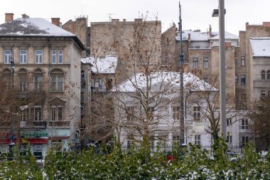 Şehir manzarasında çatılar ve ağaç dalları karla kaplanır. İnsanlar yanından geçer ve binalar tipik bir kış gününde birbirlerine yakın dururlar. Ön planda bir park görülebilir..