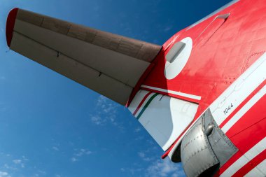 Açık mavi gökyüzüne karşı kırmızı ve beyaz bir JAK-40E uçak kanadı gösteriliyor. Uçağın kuyruğu öne çıkıyor ve hava açık ve güneşli görünüyor. Manzara havaalanında bir anı yakalıyor..