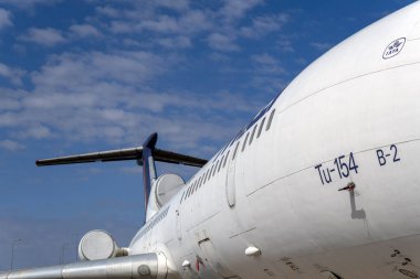 Eski bir Tu-154 uçağı havaalanında hareketsiz duruyor. Uçak, mavi gökyüzüne karşı gövdesini ve kuyruğunu gösteren bir açıdan gösterilir. Olay yeri, uçağın detaylarını yakalıyor..