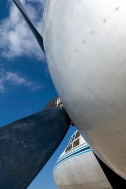 Açık mavi gökyüzünde bulutlu bir uçak pervanesinin yakın görüntüsü. Sahne motor bileşenlerini düşük açılı perspektiften gösterir.