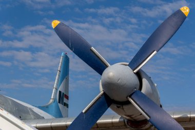 Bir uçak pervanesinin yakından görüntüsü detaylarını mavi gökyüzüne karşı gösterir. Uçağın kuyruğu görünüyor, park halinde olduğunu gösteriyor. Arkaplanda bulutlar sürükleniyor.
