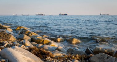 Taş sahil kıyısı, dalgalar ve kargo gemileri arka plandan limana girmek için sırada bekliyorlar.