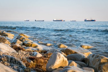 Taş sahil kıyısı, dalgalar ve kargo gemileri arka plandan limana girmek için sırada bekliyorlar.