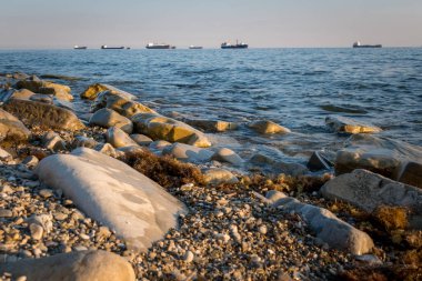 Taş sahil kıyısı, dalgalar ve kargo gemileri arka plandan limana girmek için sırada bekliyorlar.