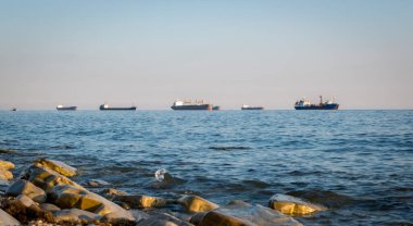 Taş sahil kıyısı, dalgalar ve kargo gemileri arka plandan limana girmek için sırada bekliyorlar.