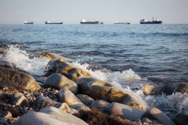 Taş sahil kıyısı, dalgalar ve kargo gemileri arka plandan limana girmek için sırada bekliyorlar.