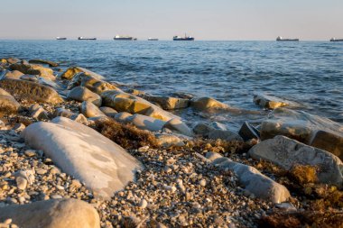 Taş sahil kıyısı, dalgalar ve kargo gemileri arka plandan limana girmek için sırada bekliyorlar.