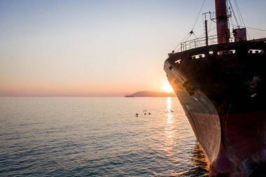 Büyük bir kargo gemisi karaya oturmuş. Çamurlu deniz kıyısında gün batımı. Günbatımı arka planında SUP tahtalarındaki insanların siluetleri