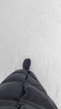 Kışın soğuk ve bulutlu bir parkta beyaz bir kar yığınının üzerinde yürüyen uzun bir kışlık ceket giyen biri. Yukarıdan bak, ayaklarına bak.