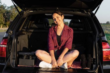 Mutlu güzel kız arabanın bagajında oturuyor ve yol kenarında günbatımının tadını çıkarıyor. Arabadaki genç bayan.