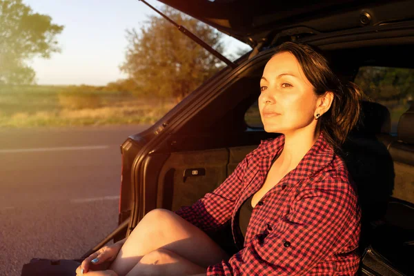 車のトランクに座って道端で夕日を楽しむ幸せな美しい女の子 車の中の若い女性は — ストック写真