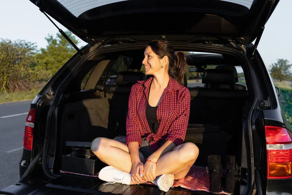 車のトランクに座って道端で夕日を楽しむ幸せな美しい女の子 車の中の若い女性は — ストック写真